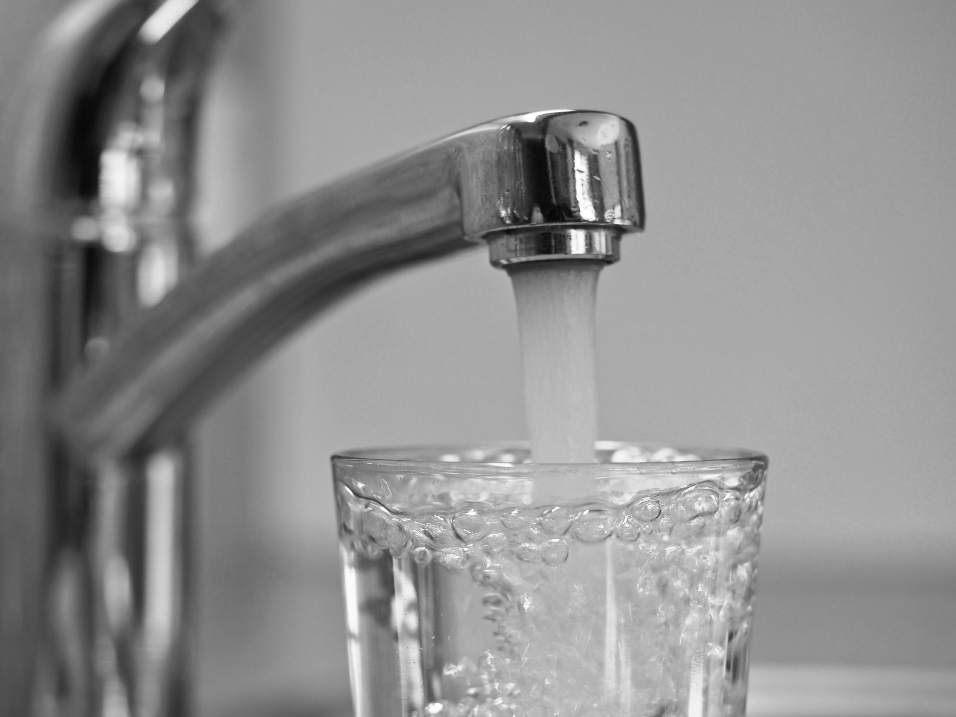 Nahaufnahme eines geöffneten Wasserhahns, aus dem Wasser in ein darunter stehendes Trinkglas fließt. Das Glas ist bereits weitgehend mit klarem Wasser gefüllt.