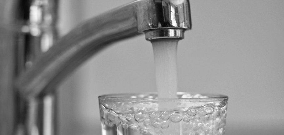 Nahaufnahme eines geöffneten Wasserhahns, aus dem Wasser in ein darunter stehendes Trinkglas fließt. Das Glas ist bereits weitgehend mit klarem Wasser gefüllt.