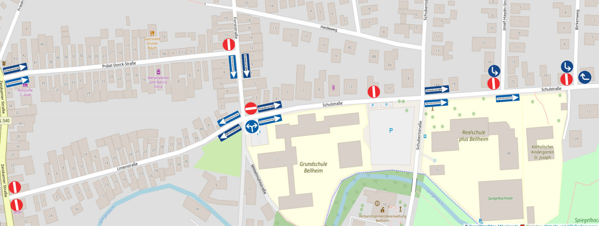 Kartenausschnitt von Bellheim mit Darstellung einer geänderten Verkehrsführung im Bereich der Grundschule Bellheim und der Realschule plus Bellheim. Betroffen sind insbesondere die Schulstraße, die Prälat-Stock-Straße sowie die Forststraße. Blaue Pfeile und Einbahnstraßenschilder kennzeichnen die vorgeschriebenen Fahrtrichtungen, rote Verkehrszeichen markieren gesperrte Zufahrten. Die Schulstraße ist abschnittsweise als Einbahnstraße ausgewiesen. Weitere Verkehrszeichen zeigen vorgeschriebene Abbiegerichtungen an den Kreuzungen. Zur Orientierung sind unter anderem die Grundschule Bellheim, die Realschule plus Bellheim, der katholische Kindergarten St. Joseph, die Spiegelbachhalle, die Verbandsgemeindeverwaltung Bellheim sowie umliegende Straßen und Gebäude eingezeichnet.
