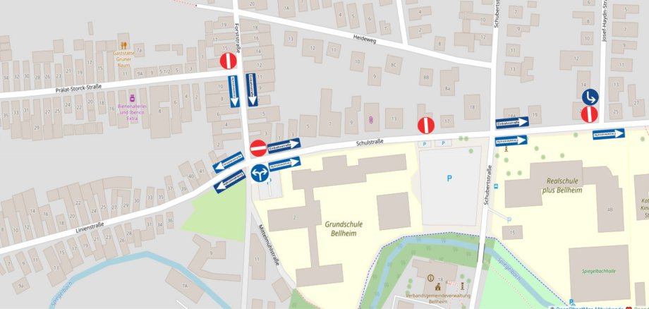 Kartenausschnitt von Bellheim mit Darstellung einer geänderten Verkehrsführung im Bereich der Grundschule Bellheim und der Realschule plus Bellheim. Betroffen sind insbesondere die Schulstraße, die Prälat-Stock-Straße sowie die Forststraße. Blaue Pfeile und Einbahnstraßenschilder kennzeichnen die vorgeschriebenen Fahrtrichtungen, rote Verkehrszeichen markieren gesperrte Zufahrten. Die Schulstraße ist abschnittsweise als Einbahnstraße ausgewiesen. Weitere Verkehrszeichen zeigen vorgeschriebene Abbiegerichtungen an den Kreuzungen. Zur Orientierung sind unter anderem die Grundschule Bellheim, die Realschule plus Bellheim, der katholische Kindergarten St. Joseph, die Spiegelbachhalle, die Verbandsgemeindeverwaltung Bellheim sowie umliegende Straßen und Gebäude eingezeichnet.