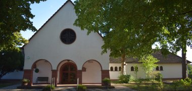 Frontansicht der Friedhofskapelle mit rundem Fenster, überdachtem Eingangsbereich und umgebenden Bäumen.