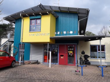 Eingangsbereich der Kindertagesstätte Spatzennest mit gelb-blauer Fassade, roter Eingangstür und der Aufschrift „Spatzennest“.