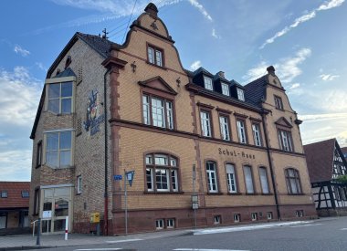 Außenansicht des Bürgerhauses Ottersheim, eines historischen, mehrgeschossigen Backsteingebäudes mit der Aufschrift „Schul-Haus“ an der Fassade. Das Gebäude besitzt eine gelbliche Backsteinfassade mit rötlichen Sandsteinelementen, hohen Fenstern und zwei markanten geschwungenen Ziergiebeln. Im Dach befinden sich mehrere Gauben. An der linken Gebäudeseite ist der Eingang mit einer Beschilderung des Malteser Hilfsdienstes zu sehen. Vor dem Gebäude verlaufen eine Straße und ein Gehweg mit Bushaltestelle. Rechts schließt ein Fachwerkgebäude an. Im Hintergrund ist ein blauer, leicht bewölkter Himmel zu sehen.