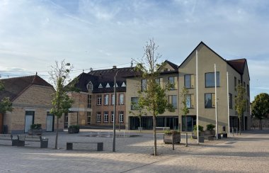 Ansicht des Dorfplatzes in Ottersheim mit einer großzügigen gepflasterten Platzfläche. Im Hintergrund stehen mehrere Gebäude, die historische Backsteinarchitektur mit moderner Bauweise verbinden. Rechts befindet sich ein mehrgeschossiges Gebäude mit heller Fassade, großen Fenstern und markanten Giebeldächern. Der Platz ist mit jungen Bäumen, Pflanzkübeln, Sitzbänken und moderner Straßenbeleuchtung gestaltet. In der Mitte befindet sich ein ebenerdiges Wasserspiel mit mehreren kleinen Fontänen. Der weitläufige, offen gestaltete Dorfplatz ist überwiegend gepflastert und wird von den umliegenden Gebäuden eingerahmt.