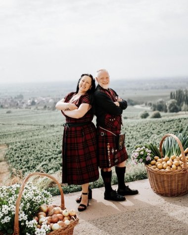 Das Zäskemmer Zwiebelkönigspaar Jürgen der Erste und Tatjana die Erste in rot-schwarz karierter Kleidung vor einer Feldlandschaft, umgeben von Körben mit Zwiebeln, Kartoffeln und Blumen.