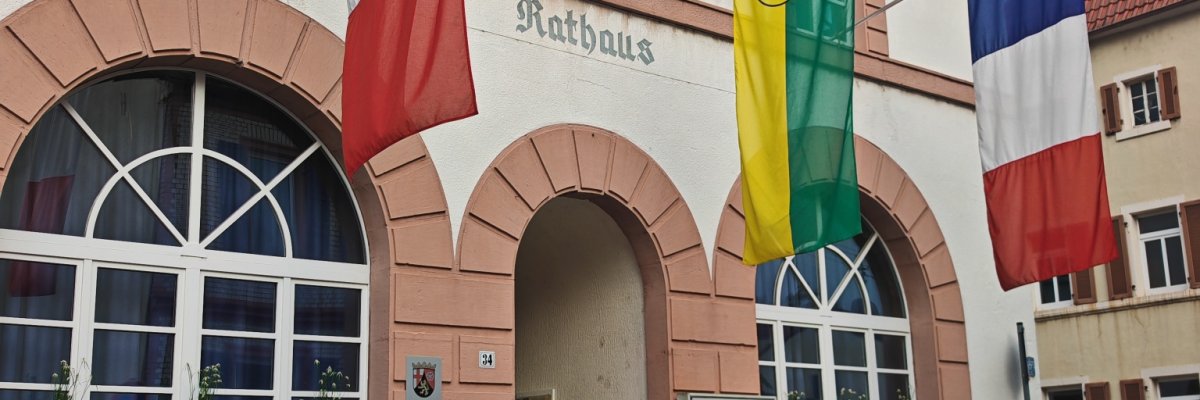 Außenansicht des historischen Rathauses in Zeiskam mit seiner markanten Fassade und Rundbogenfenstern. Vor dem Gebäude wehen die Flaggen von Italien, Zeiskam und Frankreich.