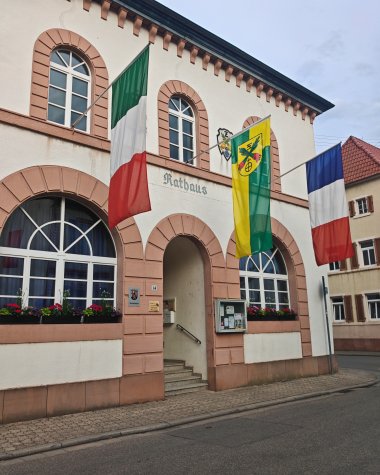 Außenansicht des historischen Rathauses in Zeiskam mit seiner markanten Fassade und Rundbogenfenstern. Vor dem Gebäude wehen die Flaggen von Italien, Zeiskam und Frankreich.