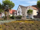Auf dem Lordplatz in Bellheim steht eine steinerne Figur mit Hut und Schnurrbart in einem blühenden Beet. Dahinter sind Bäume, Häuser und ein geparktes Auto zu sehen.