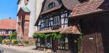Kirche im Hintergrund, davor ein Fachwerkhaus mit bewachsener Fassade. Im Hintergrund strahlend blauer Himmel