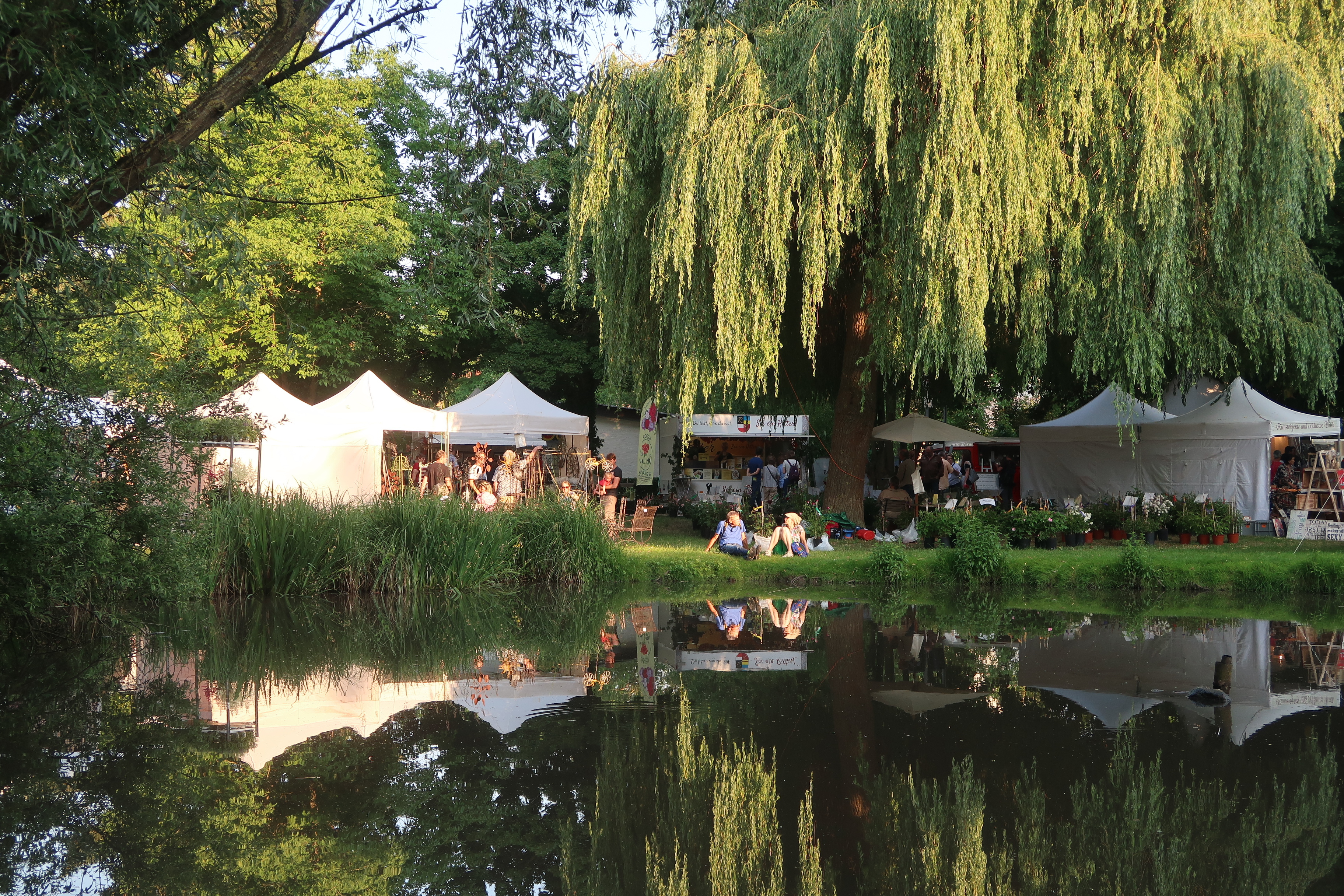 Ein Veranstaltungsfoto der VG Bellheim mit mehreren Personen bei einer Festlichkeit, den Bellheimern Gartentagen. Zu sehen Wasser (Teich), Festzelte und Menschen stehend und sitzend auf Bänken