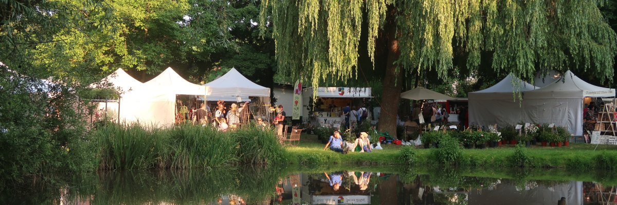 Ein Veranstaltungsfoto der VG Bellheim mit mehreren Personen bei einer Festlichkeit, den Bellheimern Gartentagen. Zu sehen Wasser (Teich), Festzelte und Menschen stehend und sitzend auf Bänken
