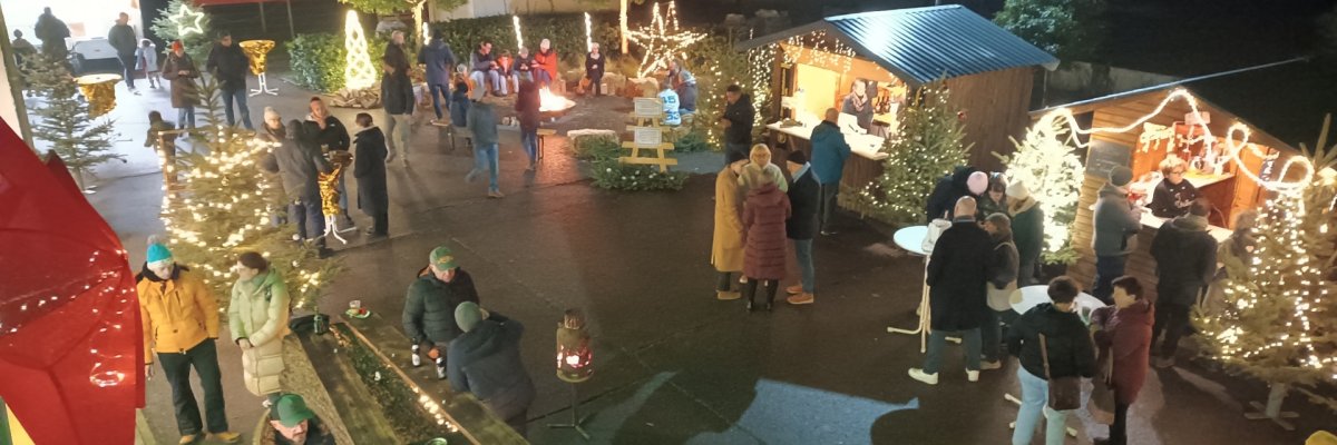 Blick auf den weihnachtlich beleuchteten Christkindlmarkt in Knittelsheim mit zahlreichen Besucherinnen und Besuchern, geschmückten Tannenbäumen, Holzhütten, Lichterketten und einer Feuerstelle.