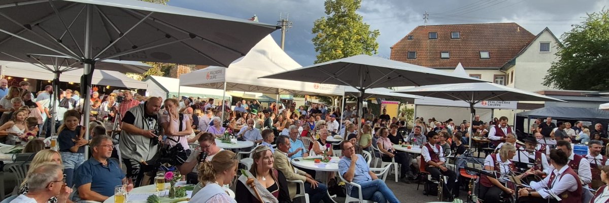 Kirwe Zeiskam Zahlreiche Menschen sitzen bei der gut besuchten Kirwe unter großen Sonnenschirmen und Festzelten. Rechts spielt eine Blaskapelle, während dunkle Woklen über dem Festplatz aufziehen