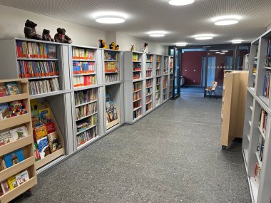 Innenansicht der Bücherei Ottersheim. Zu beiden Seiten eines breiten Ganges stehen zahlreiche Bücherregale mit einer großen Auswahl an Büchern, darunter viele Kinder- und Jugendbücher. Auf der linken Seite befinden sich zusätzliche Präsentationsregale, in denen Bücher mit sichtbaren Covern ausgestellt sind. Auf den Regalen stehen mehrere Stofftiere und Figuren als Dekoration. Der Raum ist modern und hell gestaltet, mit grauem Fliesenboden, weißen Wänden und großen runden Deckenleuchten. Im hinteren Bereich führen Glastüren in einen angrenzenden Raum.