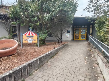 Außenansicht des Gemeindekindergartens „Flohzirkus“ mit gepflastertem Zugangsweg, Bäumen und dem farbig gestalteten Eingangsschild vor dem Gebäude.