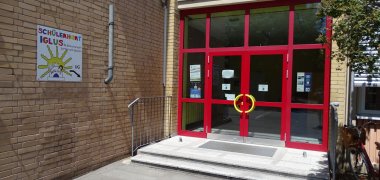 Eingangsbereich des Schülerhorts „Iglus“ mit roter Glasfront und gelben Türgriffen. Links weist ein bunt gestaltetes Schild auf den Schülerhort hin.