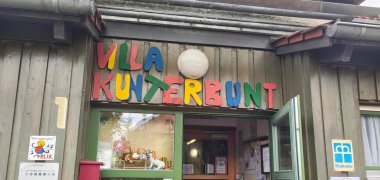 Eingangsbereich der Kindertagesstätte „Villa Kunterbunt“ in einem Holzgebäude. Über der geöffneten Eingangstür befindet sich der Name der Einrichtung in großen, bunten Buchstaben.
