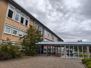 Außenansicht der Grundschule Bellheim mit mehrgeschossigem Schulgebäude, zahlreichen Fensterreihen und einem verglasten Eingangsbereich mit roten Türen. Im Vordergrund befinden sich der gepflasterte Schulhof sowie Bäume und Sträucher.